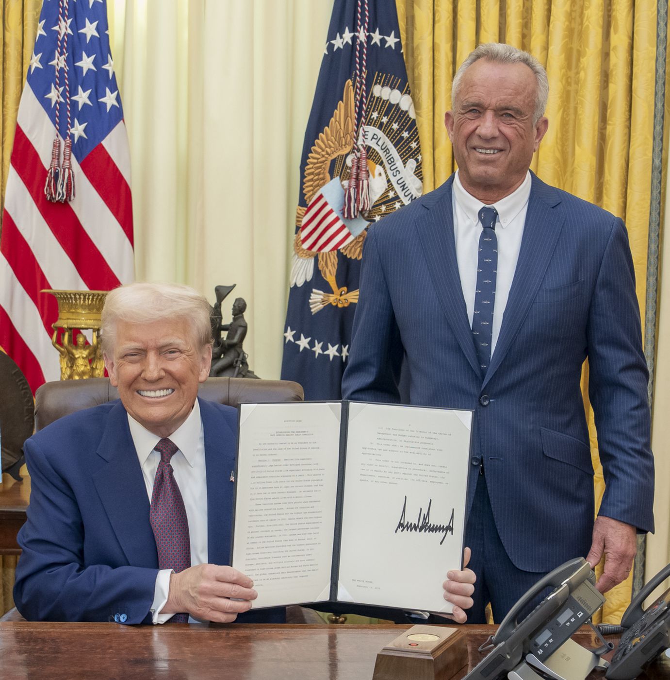 President Trump, seated at the Resolute Desk, holds Secretary Kennedy’s signed commission as Kennedy stands beside him